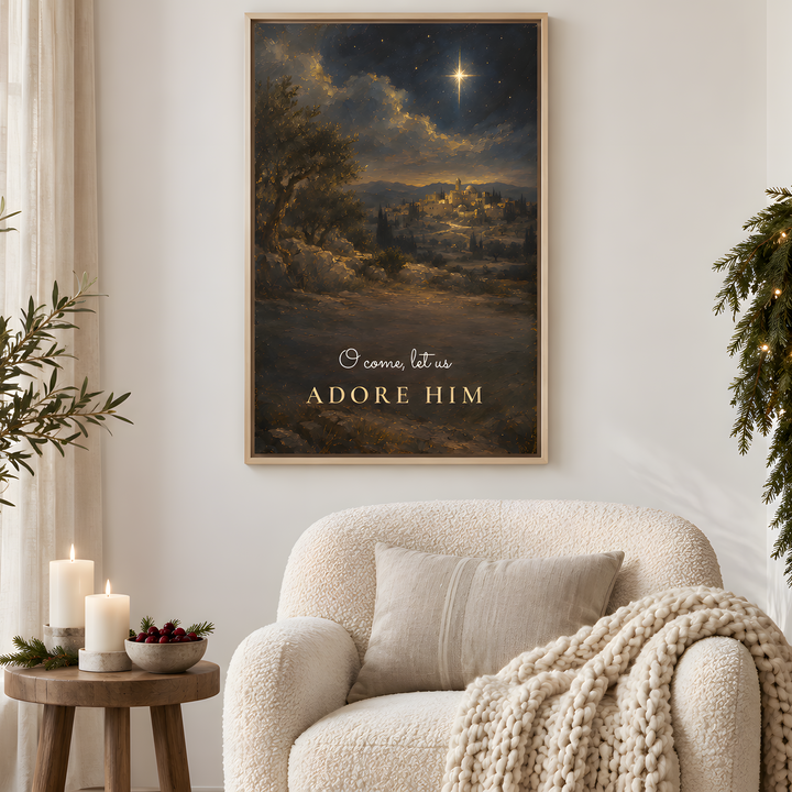 A framed art print featuring a Christmas night scene with the text 'Come let us adore him' hangs on a white wall above a cozy cream-colored armchair in a decorated living room.