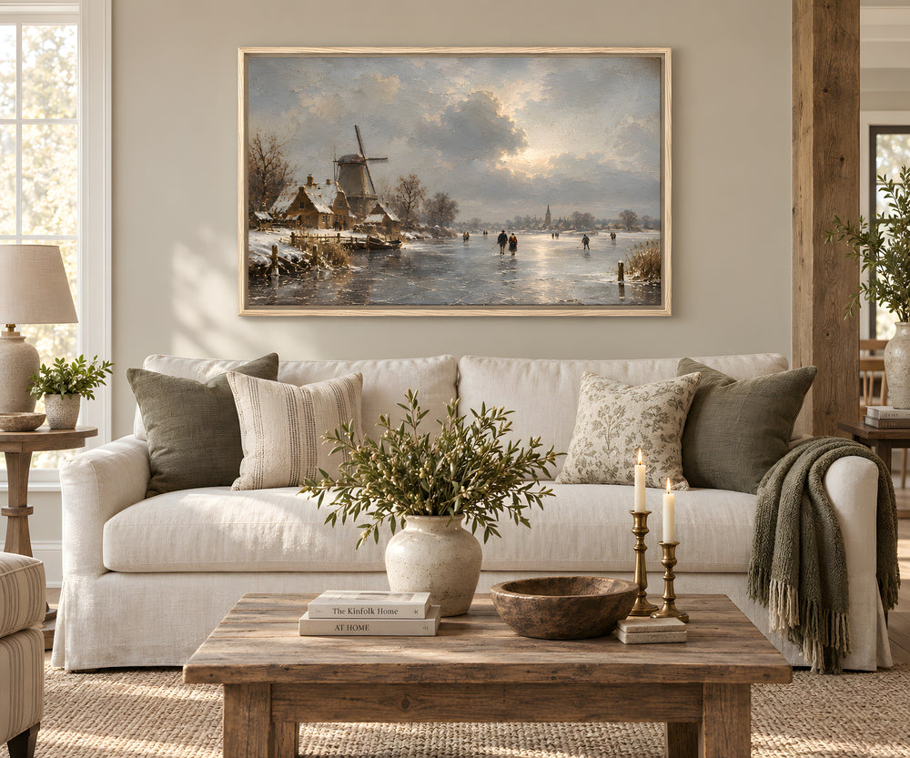 A framed landscape painting of a Dutch winter scene with a windmill, ducks, and figures on the ice, displayed above a white sofa in a styled living room.