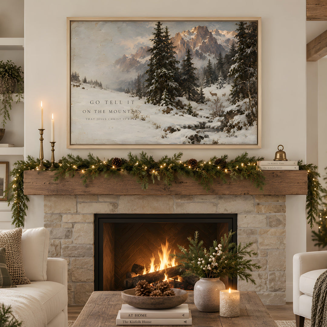 A large framed winter landscape painting hangs above a stone fireplace in a cozy living room. The artwork shows snow-covered pine trees and mountains with the text 'GO TELL IT ON THE MOUNTAIN.