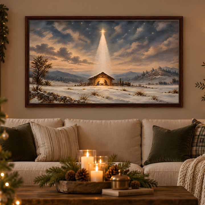 A framed Christmas landscape painting hangs above a stone fireplace in a cozy living room. The painting depicts a snowy winter scene with a small stable under a glowing star in the night sky.