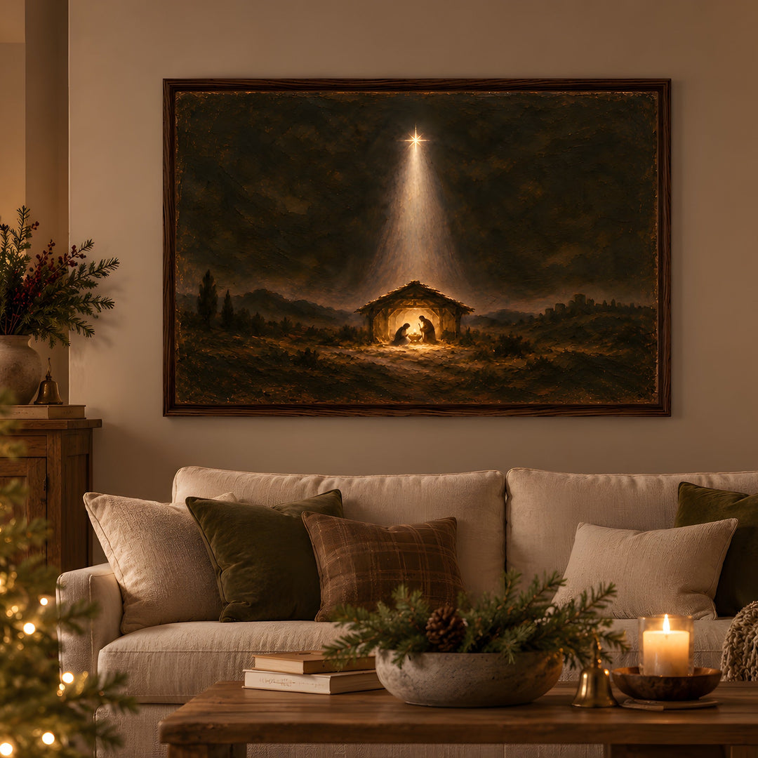 A framed Christmas nativity painting hangs on a beige wall above a white sofa in a cozy living room. The painting depicts a dark night scene with a glowing light beam over a stable where a baby and Mary sit.