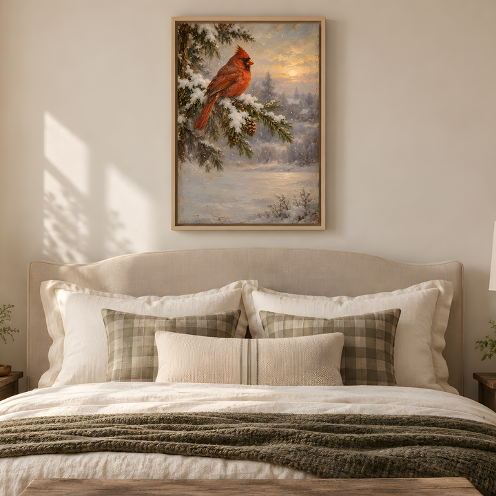 A framed art print of a red cardinal perched on a snowy pine branch, hanging on a beige wall above a neatly made bed with white and plaid pillows in a warm, rustic bedroom setting.