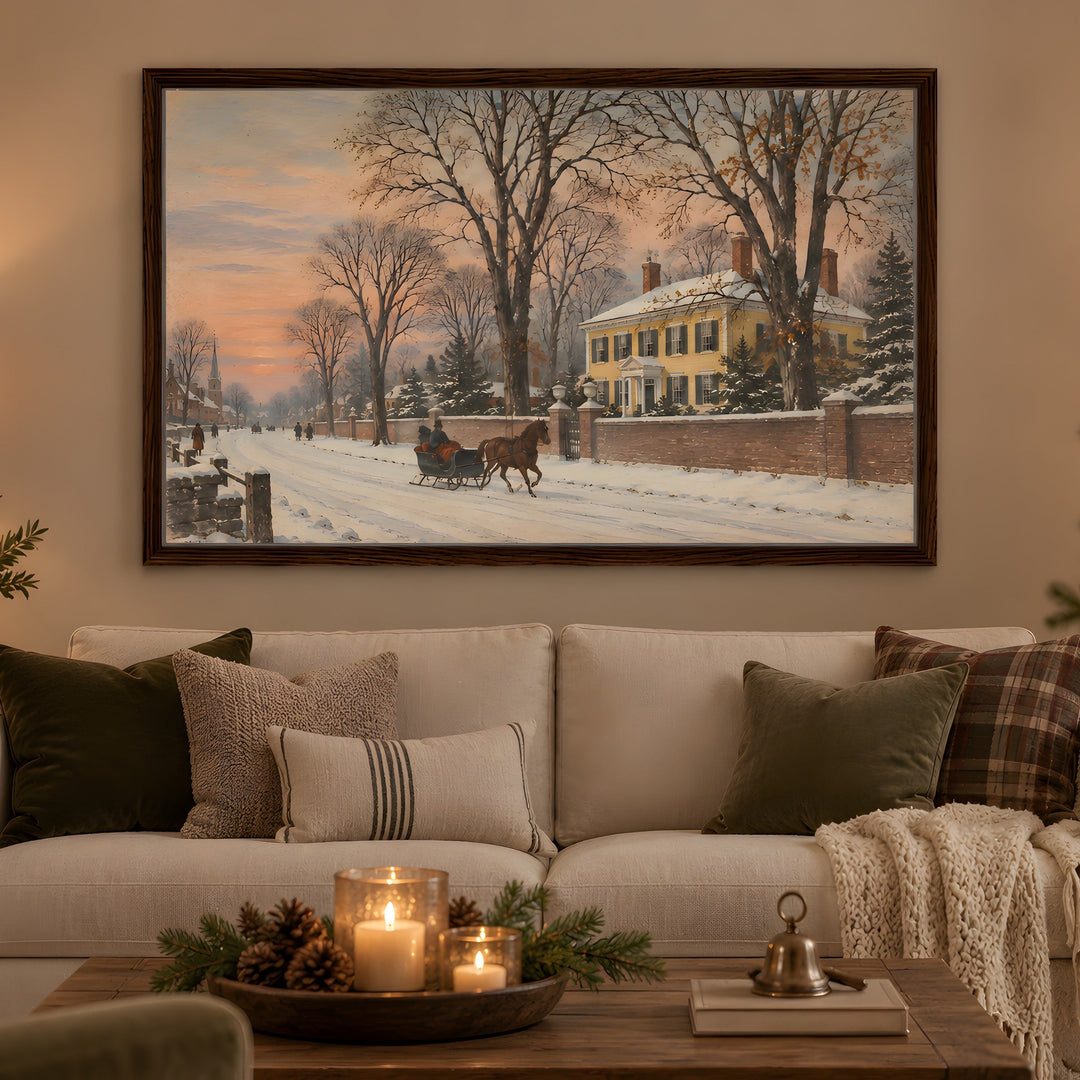 A framed winter landscape painting hangs on a beige wall above a white sofa in a cozy living room. The painting depicts a snowy street scene with bare trees, a yellow house, and a horse-drawn carriage.