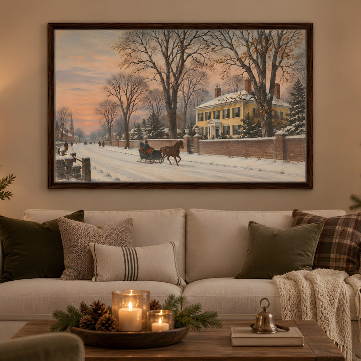 A framed winter landscape painting hangs on a beige wall above a white sofa in a cozy living room. The painting depicts a snowy street scene with bare trees, a yellow house, and a horse-drawn carriage.