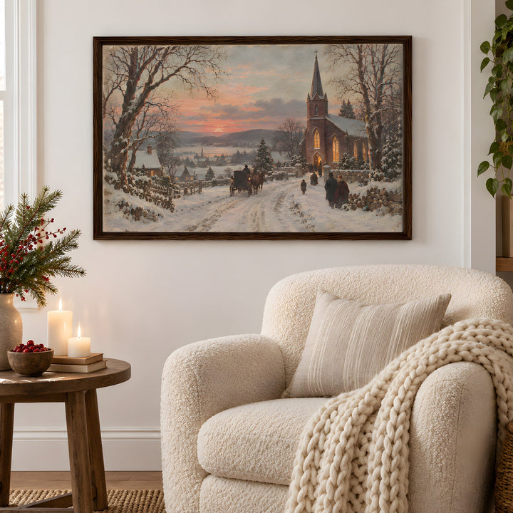 A framed winter landscape painting hangs on a white wall above a cream bouclé armchair in a cozy living room. The painting depicts a snowy village scene with a church, bare trees, and a warm sunset sky.