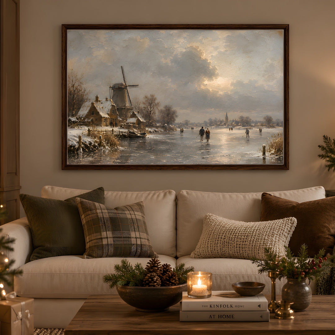 A large framed landscape painting of a snowy winter village with a windmill and ice skating scene, displayed on a beige wall above a cream sofa in a warmly lit living room with pillows, a coffee table, and holiday decor.