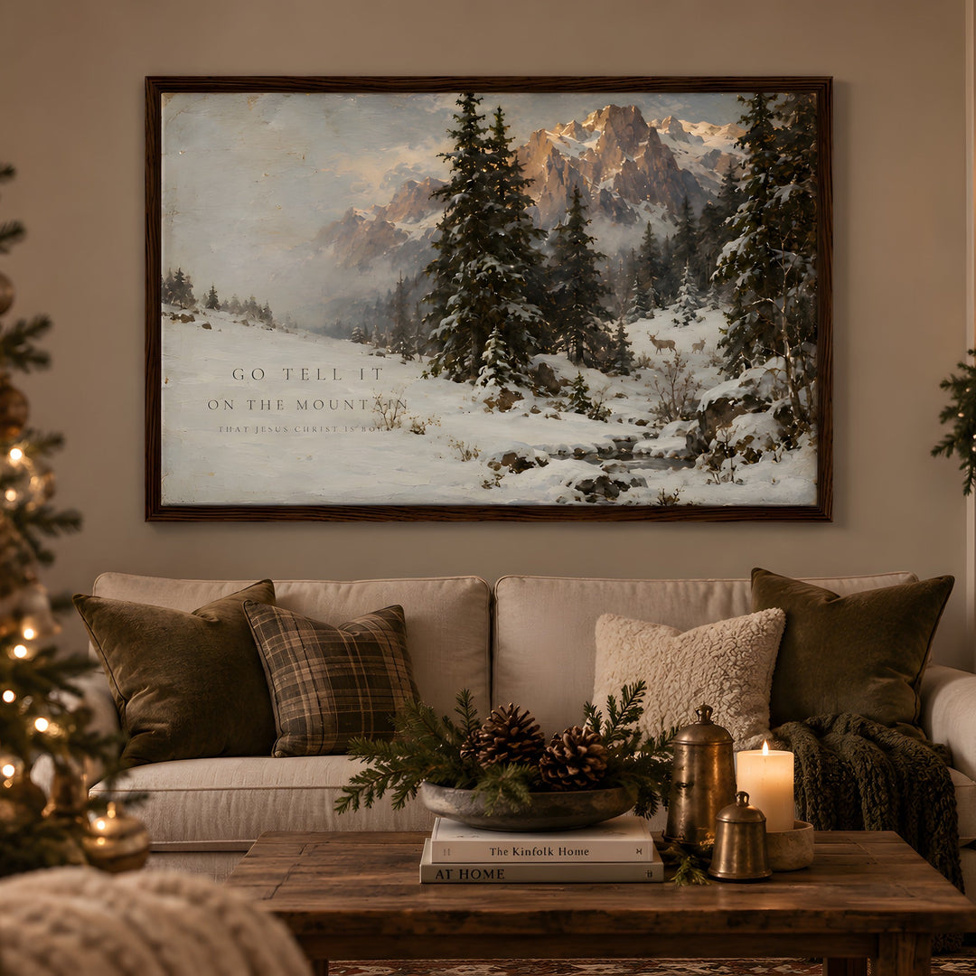 A large framed winter landscape painting hangs above a beige sofa in a cozy living room. The artwork depicts snow-covered pine trees and mountains under a soft sky, with the text 'GO TELL IT ON THE MOUNTAIN' visible in the scene.