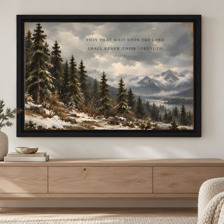 A framed landscape art print featuring a snowy pine forest and mountains with a Bible verse, displayed on a light gray wall above a wooden console table in a styled living room.