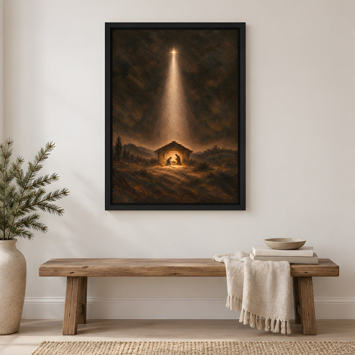 A framed vertical art print depicting a nativity scene with a bright beam of light shining down into a stable, hung on a white wall in a styled living room setting above a wooden bench.