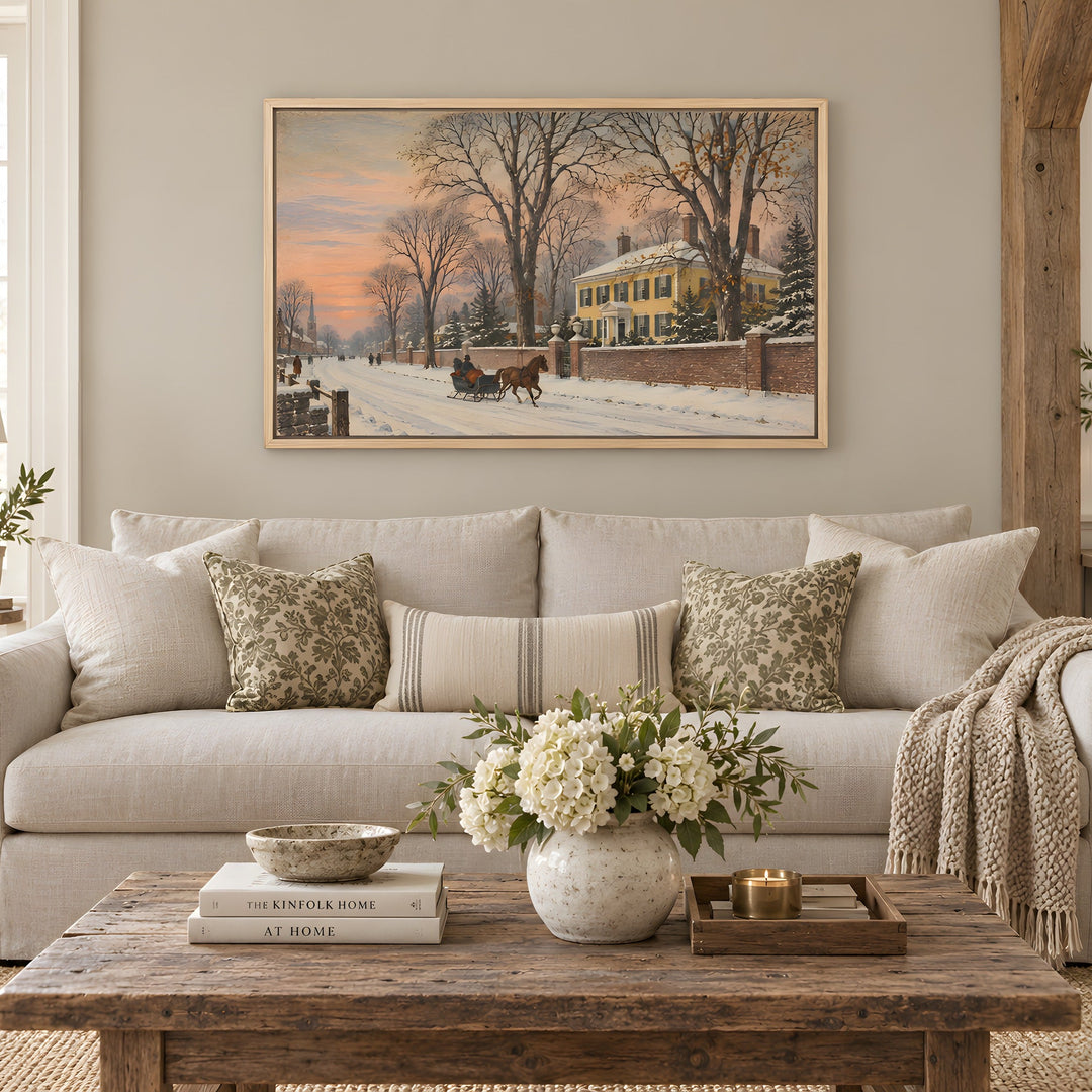 A framed winter landscape painting hangs on a beige wall above a light gray sofa in a styled living room. The painting depicts a snowy street scene with bare trees, a yellow house, and a horse-drawn carriage at sunset.