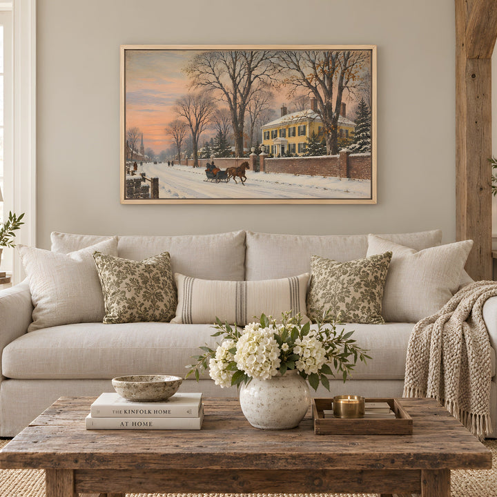 A framed winter landscape painting hangs on a beige wall above a light gray sofa in a styled living room. The painting depicts a snowy street scene with bare trees, a yellow house, and a horse-drawn carriage at sunset.