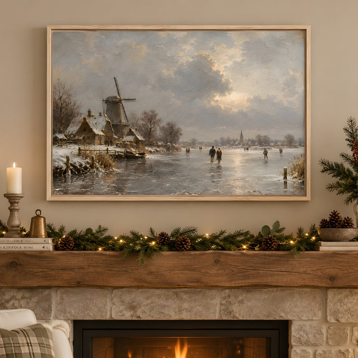 A framed landscape painting of a snowy winter village with a windmill and people skating on a frozen river, displayed above a decorated stone fireplace with candles, greenery, and pinecones.