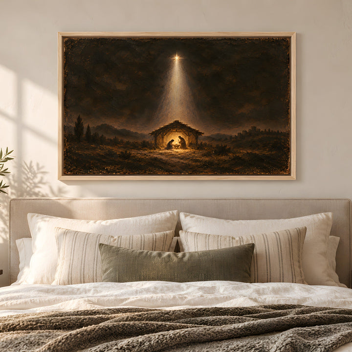 A framed dark religious art print depicting a stable under a beam of light, displayed on a beige wall above a neatly made bed with white and olive green pillows in a softly lit bedroom.