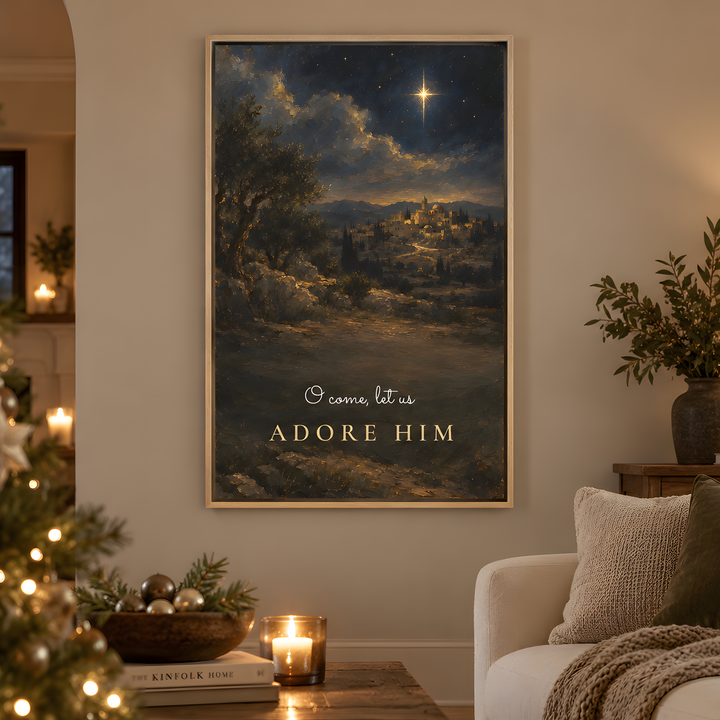 A vertical framed art print featuring a Christmas night landscape with a shining star and the text 'O come, let us ADORE HIM' in gold, displayed on a beige wall in a cozy living room setting with a lit Christmas tree, sofa, and candles.