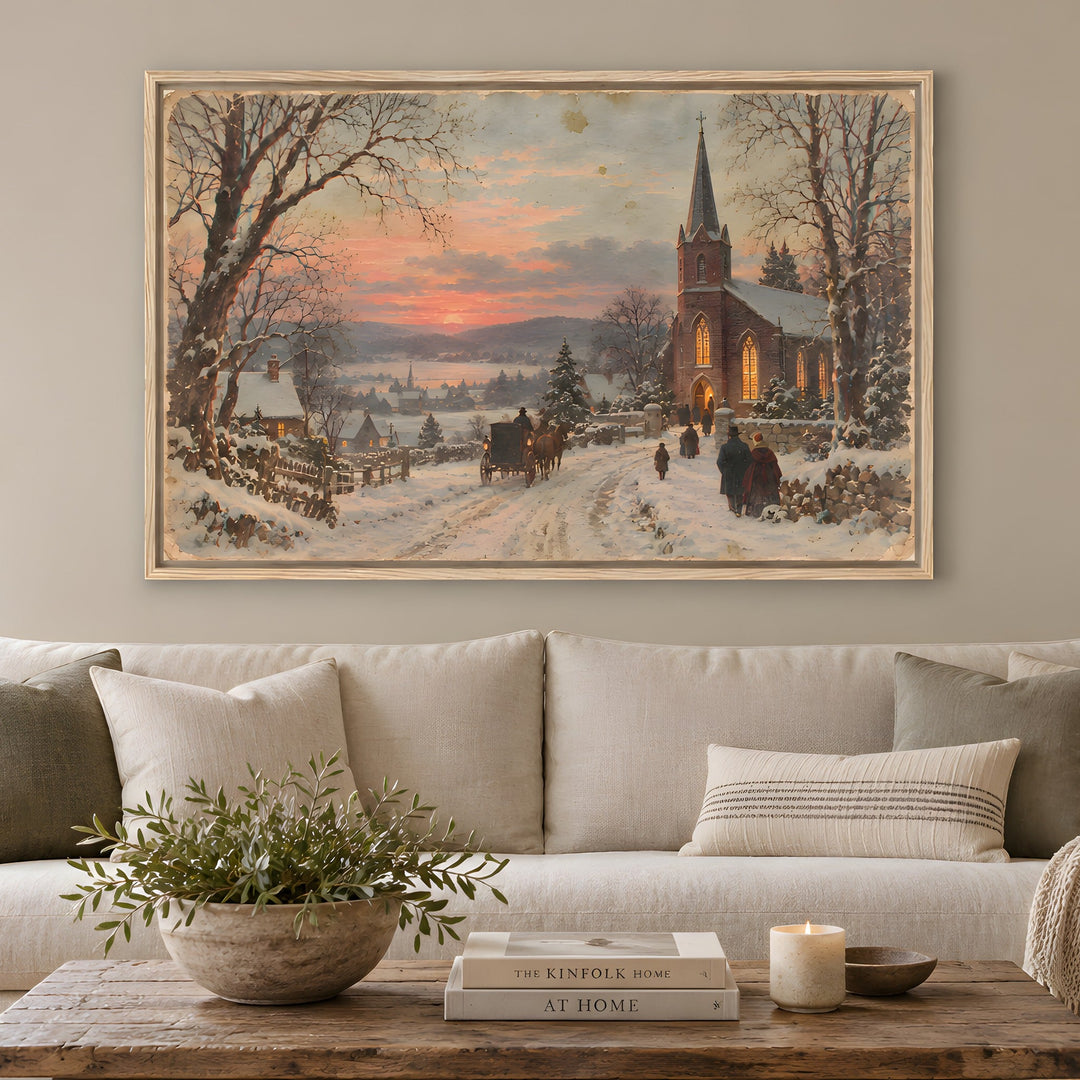 A large framed winter landscape painting hangs on a light gray wall above a cream sofa in a styled living room. The artwork depicts a snowy village street with a church, bare trees, and a warm sunset sky.