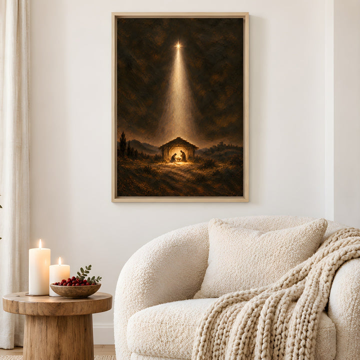 A framed vertical art print depicting a nativity scene with a bright beam of light shining down into a stable, hung on a white wall in a cozy living room setting with a cream boucle armchair, candles, and a wooden side table.
