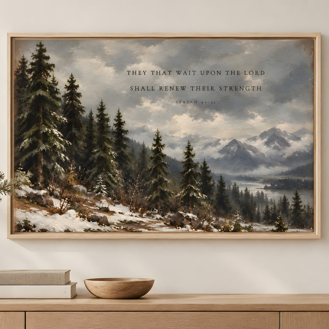 A framed landscape art print featuring a snowy pine forest and mountains with a Bible verse printed in the sky, displayed on a light wood console table with books and a bowl.