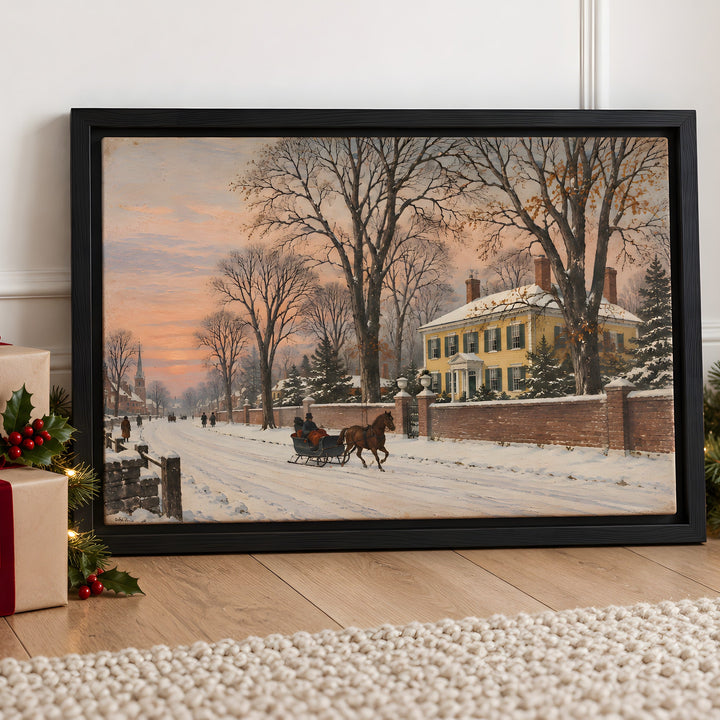 A framed winter landscape print leaning against a white wall on a wooden floor, surrounded by holiday decorations including wrapped gifts, evergreen branches, and warm string lights. The artwork shows a snowy street with bare trees, a horse-drawn sleigh, and a yellow house under a soft orange sky.