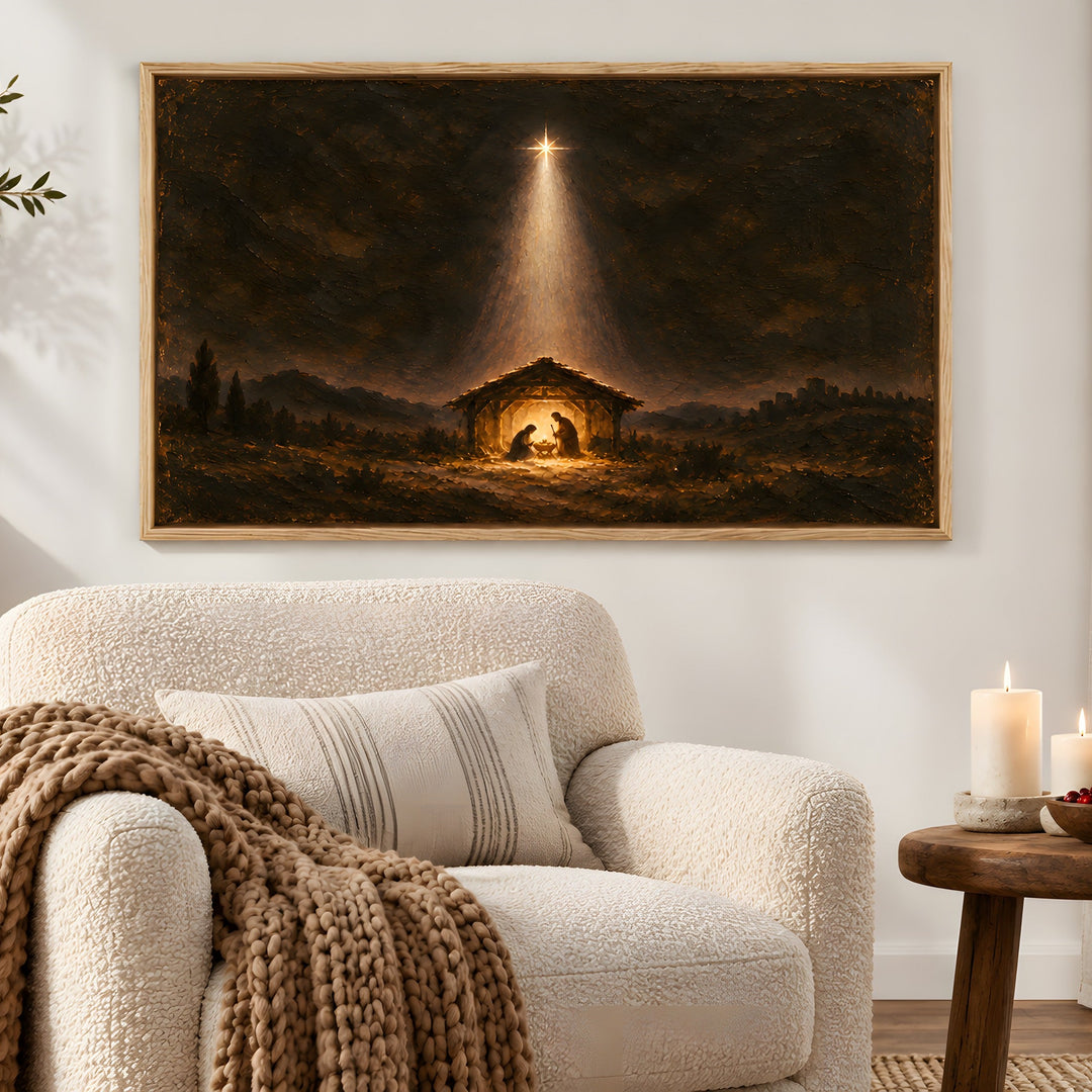 A framed religious wall art print depicting the Nativity scene with a bright star shining into a stable, hung above a cream bouclé armchair in a styled living room with candles and a side table.