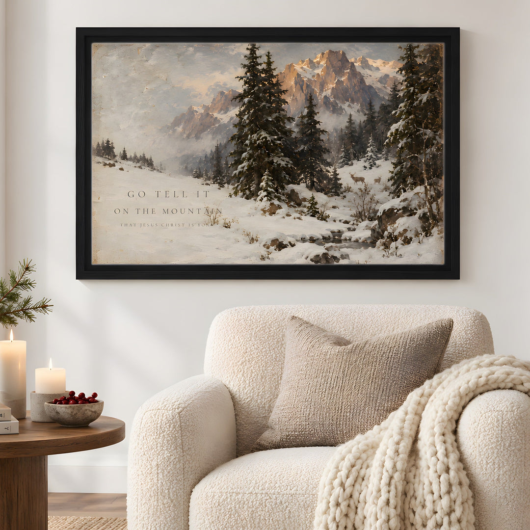 A framed winter mountain landscape print hangs on a white wall above a cream bouclé armchair with a chunky knit throw blanket. The artwork shows snow-covered evergreen trees, distant mountains, and a warm glowing peak, with small text near the lower left.