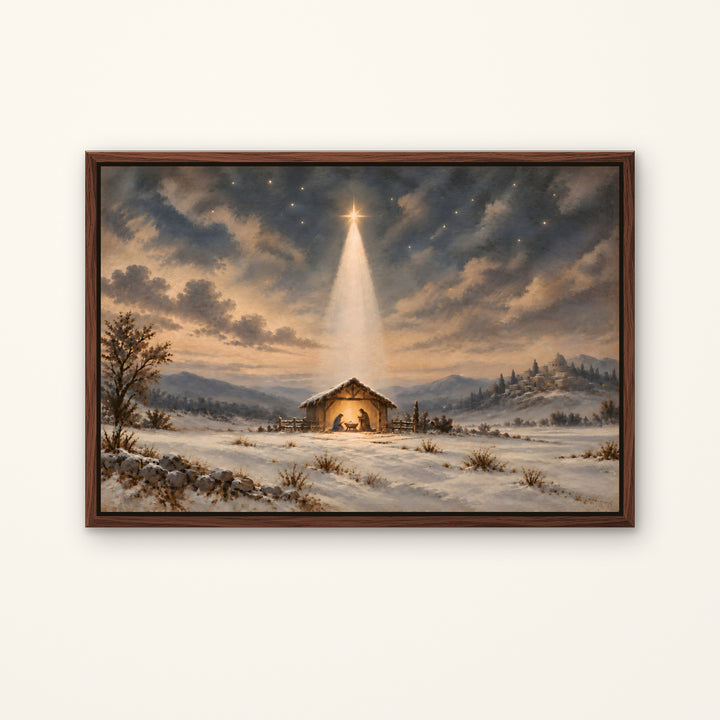 Framed wall art showing a snowy winter landscape with a small glowing stable under a dramatic cloudy sky and a bright star beam. The artwork is enclosed in a dark brown wooden frame and displayed against a light warm off-white wall.